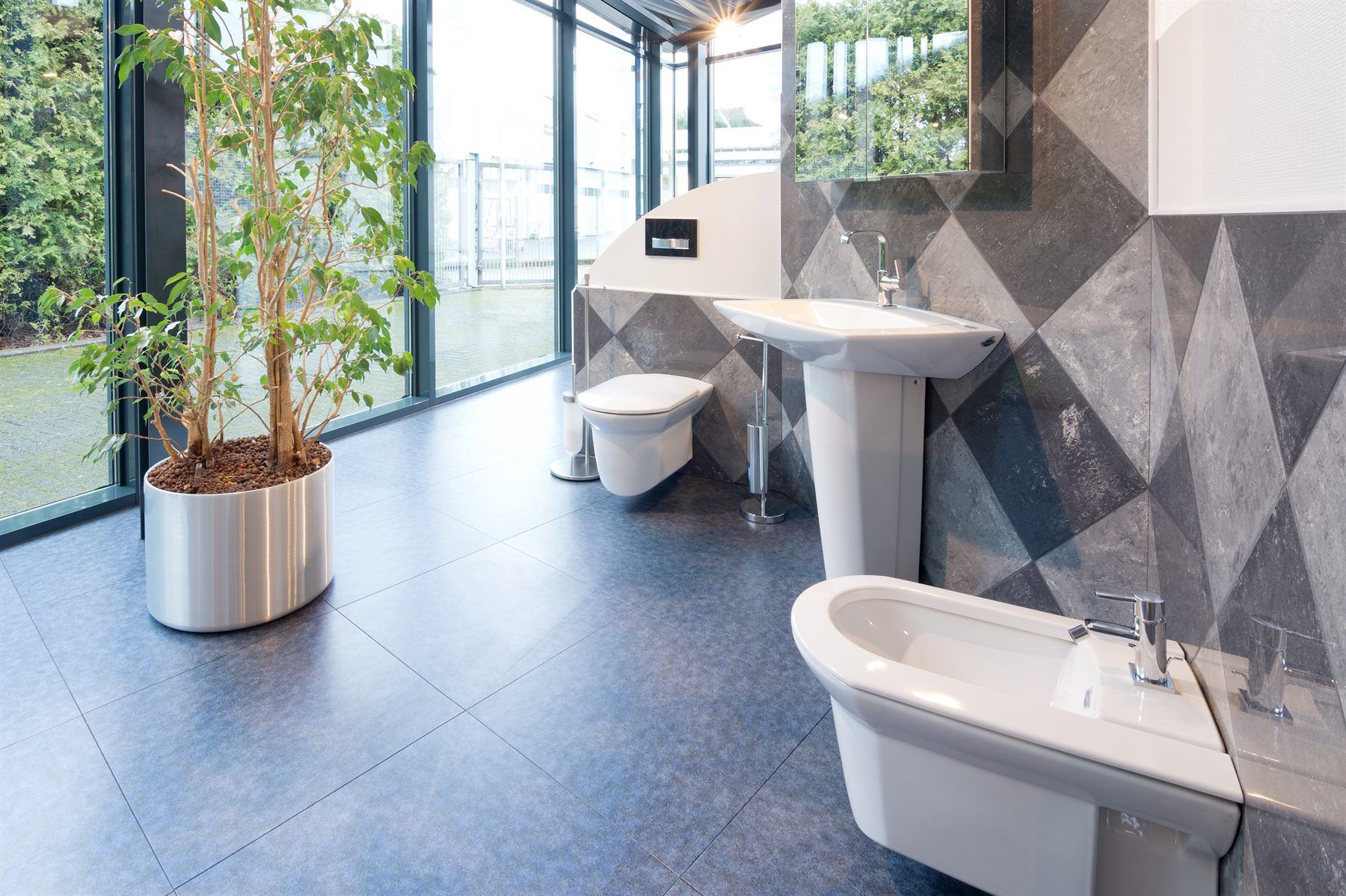 Ausstellungsbereich mit freistehender Badewanne, Waschbecken und großer Zimmerpflanze vor Glasfront
